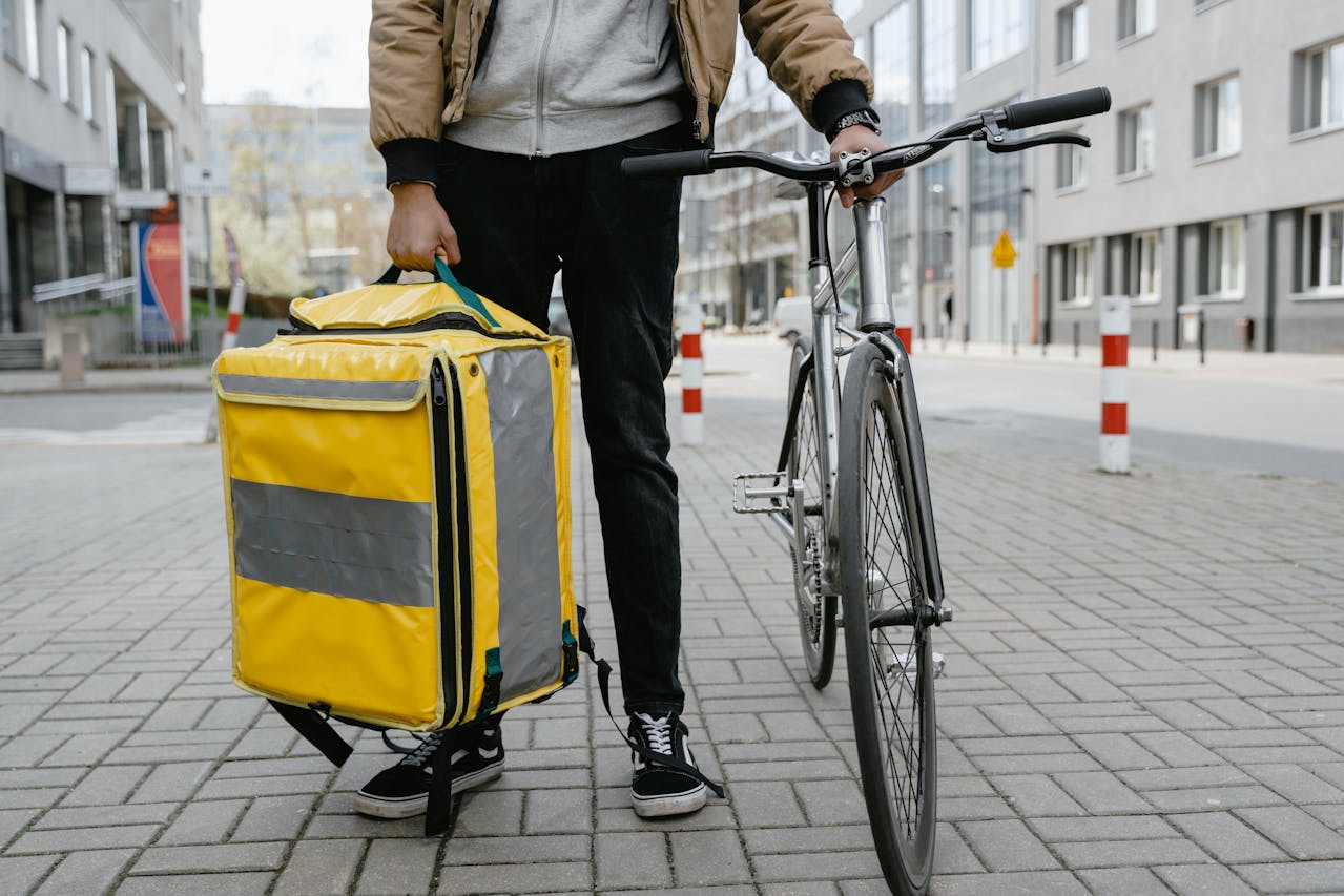 Street-level view of a person who holds a bicycle in the one hand, and a large bike delivery bag in the other. Image by Mart Production on Pexels.